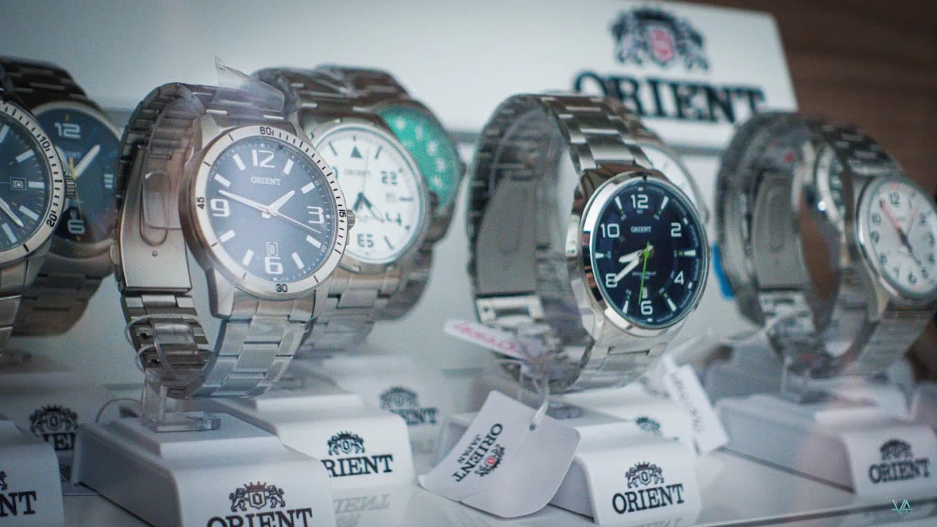 Fileira de relógios masculinos Orient de pulseira de aço, com etiquetas de papel penduradas, na vitrine iluminada